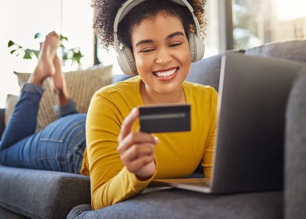 woman with headphones laying on sofa looking at credit card with laptop open